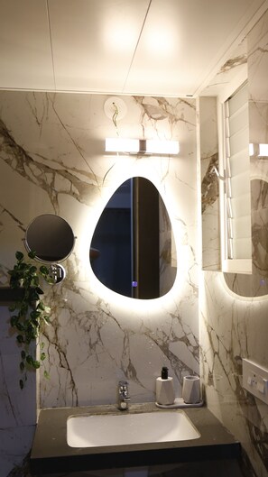 Une salle de bain avec un mur en marbre, un miroir rond, un lavabo et un luminaire placé au-dessus du miroir.