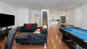 A modern living room with a pool table, a sofa, a flat-screen TV, and a staircase leading to a kitchen area.