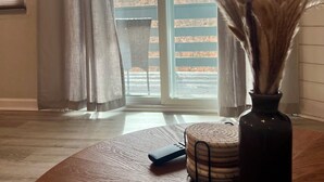 A room with a wooden floor, a round table with a vase of pampas grass, a remote control, and a window with curtains.