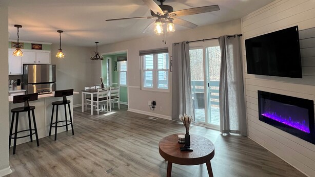 A spacious living area with a wooden floor, a ceiling fan, a TV mounted on the wall, and a fireplace.