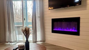 A living room with a flat-screen TV mounted on the wall, a fireplace with a purple-lit fire, and a round wooden coffee table with a vase and remote control.