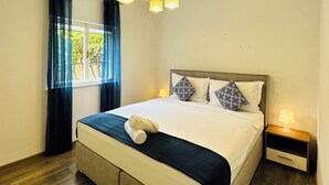 A modern hotel room with a large bed, a bedside table with a lamp, a ceiling light fixture, and a window with curtains. - BEACH HOUSE BOL (Bol)
