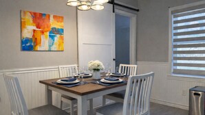 Ein Essbereich mit einem Holztisch, gedeckt für vier Personen, weißen Stühlen, einer Blumenvase und einem farbenfrohen, abstrakten Gemälde an der Wand.
