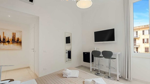 A modern hotel room with a bed, a desk with two chairs, a TV, and a large window with a city view.