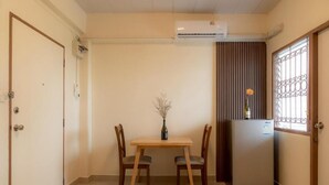 Una habitación pequeña y limpia con una mesa de madera y dos sillas, un refrigerador y una ventana con un celosía decorativa.