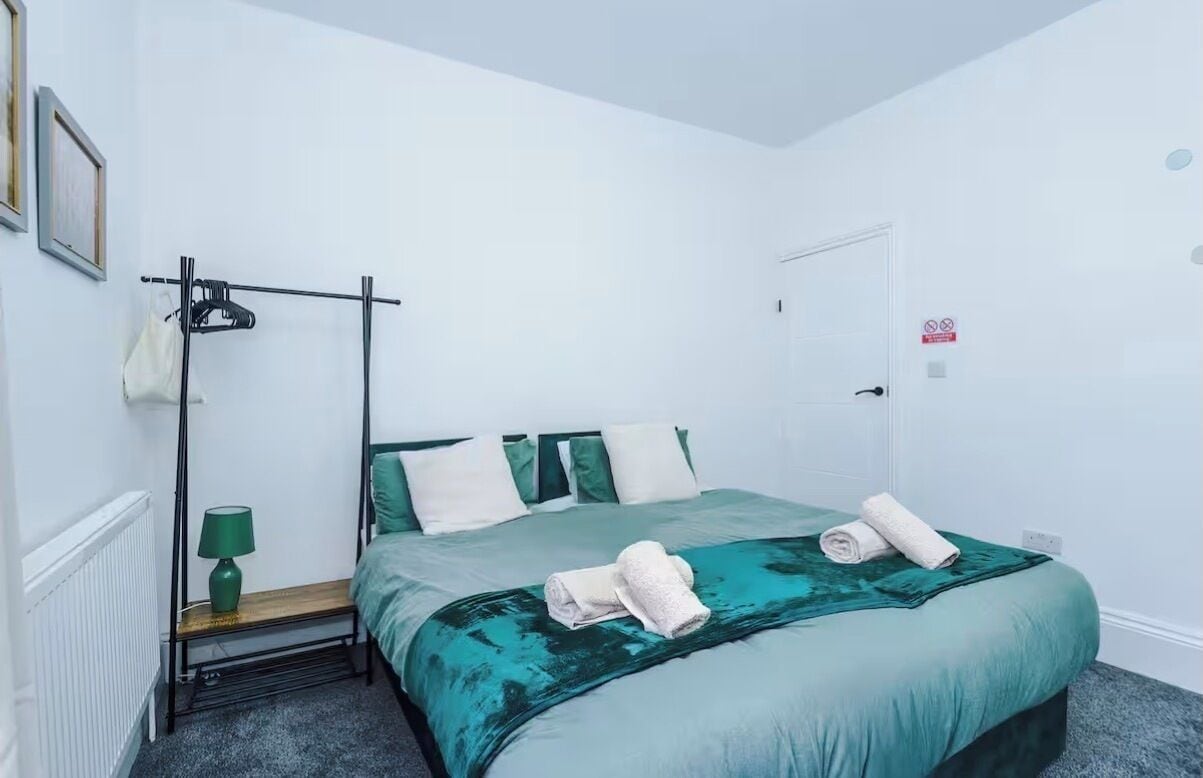 A single-bed room with a teal bedspread, a green lamp, and a black metal rack.