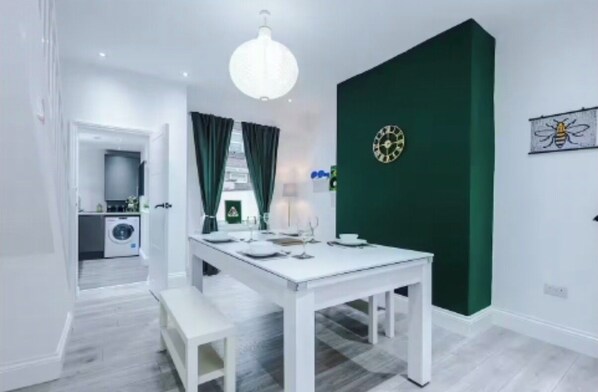Ein moderner Essbereich mit einem weißen Tisch, gedeckt für vier Personen, einer grünen Akzentwand und einer im Hintergrund sichtbaren Küche.