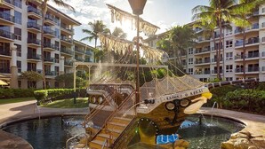 A pool area with a ship-shaped water feature, surrounded by palm trees and a multi-story building with balconies. - Cozy 1-bedroom/living room condo with AC in charming Lahaina (Lahaina)