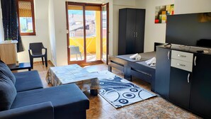 Un espacio habitable compacto con sofá, una mesita, una zona de cocina con televisor y un ventanal corredero hacia un balcón.