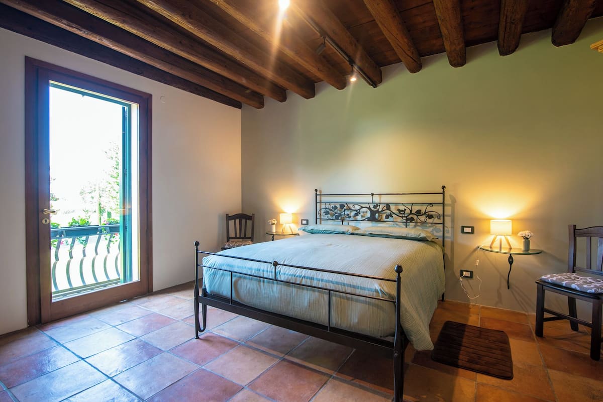 A bedroom with a bed, two bedside lamps, a wooden chair, a glass table, and a view through a door.