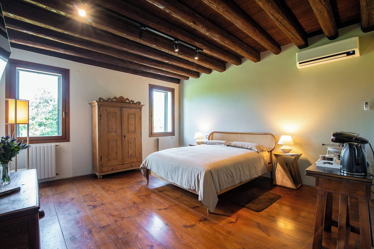 A bedroom with a wooden ceiling, a bed with a headboard, a nightstand with a lamp, a radiator, and a window.