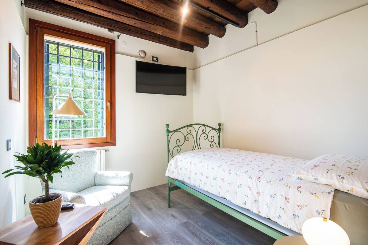 A bedroom with a bed, a window, a wall-mounted TV, and a potted plant.