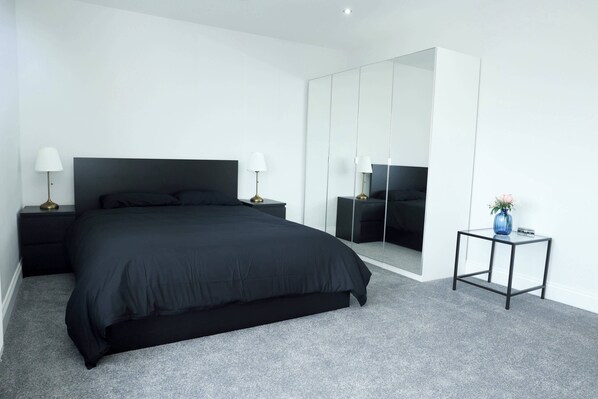 Een moderne slaapkamer met een groot bed, een garderobe, een nachtkastje met een lamp en een klein tafeltje met een vaas.