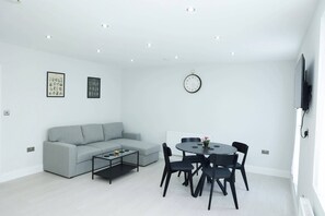 Een moderne woonkamer met een grijze bank, een salontafel, een eettafel met stoelen, een wandklok en een flatscreen televisie aan de muur.
