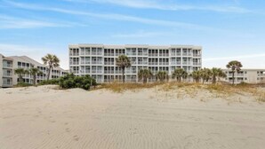 Ein Strandhotel mit mehrstöckigen Gebäuden, einem Sandstrand im Vordergrund und Palmen.