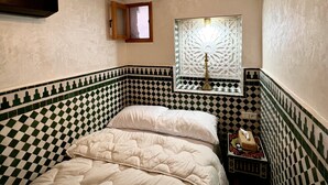 A room with a checkered floor, a bed with white bedding, a decorative ceiling, and a window with a patterned blind.