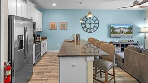 Une cuisine moderne dotée d’un îlot central, d’appareils électroménagers en acier inoxydable et d’un coin repas agrémenté d’un ventilateur de plafond et d’une horloge murale.