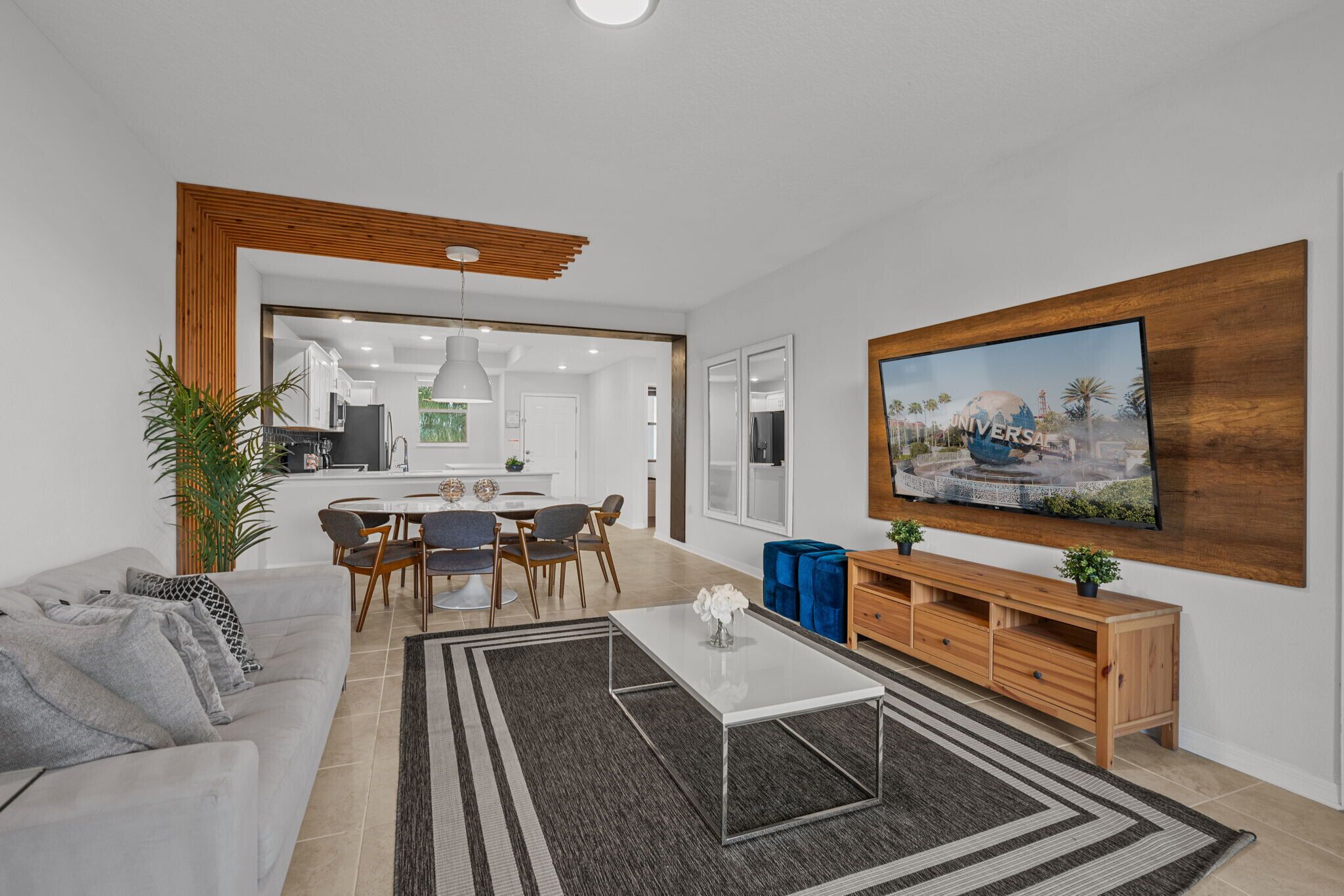 Una sala de estar moderna con un sofá, una mesa de centro y un televisor. Se aprecia un comedor con sillas y una mesa. También se observa un mueble de madera y una planta.