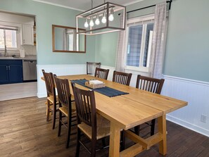 A dining room with a wooden table and chairs, a chandelier, and a kitchen in the background. - Lake Roots: Peaceful 4-bedroom lakefront home in charming Stanardsville (Stanardsville)