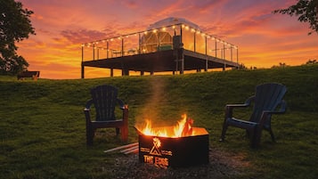 Een vuurkuil met een logo, twee Adirondack stoelen en een glazen constructie met lichtjes bij zonsondergang.