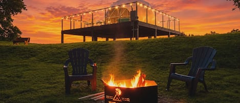 A fire pit with a logo, two Adirondack chairs, and a glass structure with string lights at sunset.
