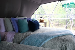 A bed with a textured blanket, pillows in various colors, and a view of greenery through a window. - Float on Inn Geodome at The Edge—Unique Riverfront Glamping in Salt Lick, KY (Salt Lick)