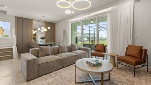Una sala moderna con un sofá modular, una mesa redonda de centro y dos sillones. Se observa un comedor al fondo, con un ventanal que da al exterior.