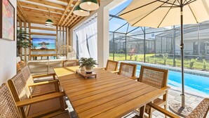 A wooden dining table with wicker chairs, a large umbrella, and a view of a swimming pool through a glass enclosure. - Last Minute Disney! Private Pool, Hot Tub, Game Room & Themed Bedrooms. Book Now! #150825 (Orlando)