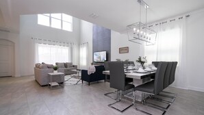 Ein modernes Wohnzimmer mit Essbereich, ausgestattet mit einem großen Fernseher, grauen Sofas und einem Chandelier.
