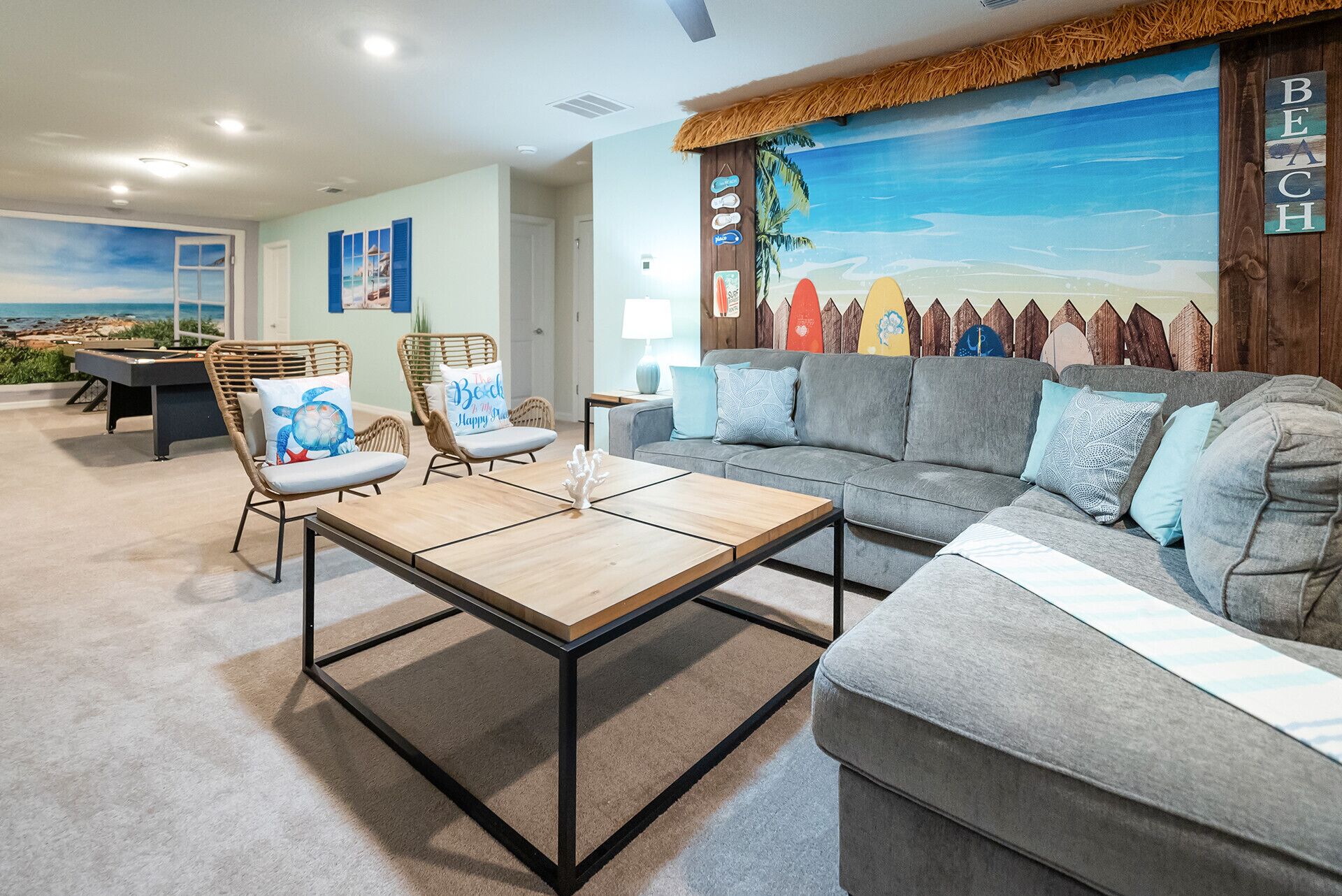 A living room with a pool table, two wicker chairs, a grey sofa, and a beach-themed mural.