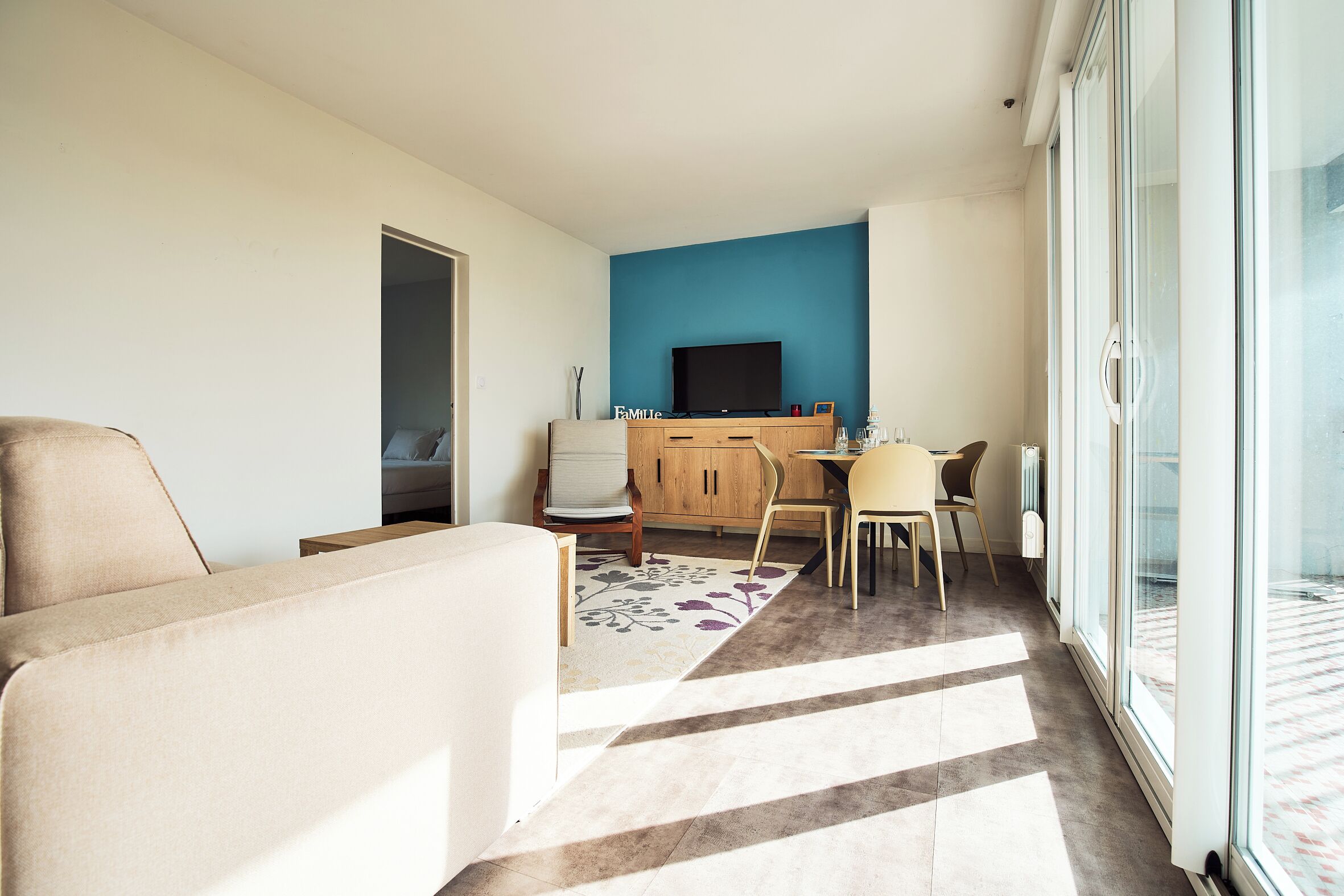 Una sala moderna con un sofá, un comedor con una mesa y sillas, un televisor en una pared azul y un amplio ventanal corredero que da a un balcón.