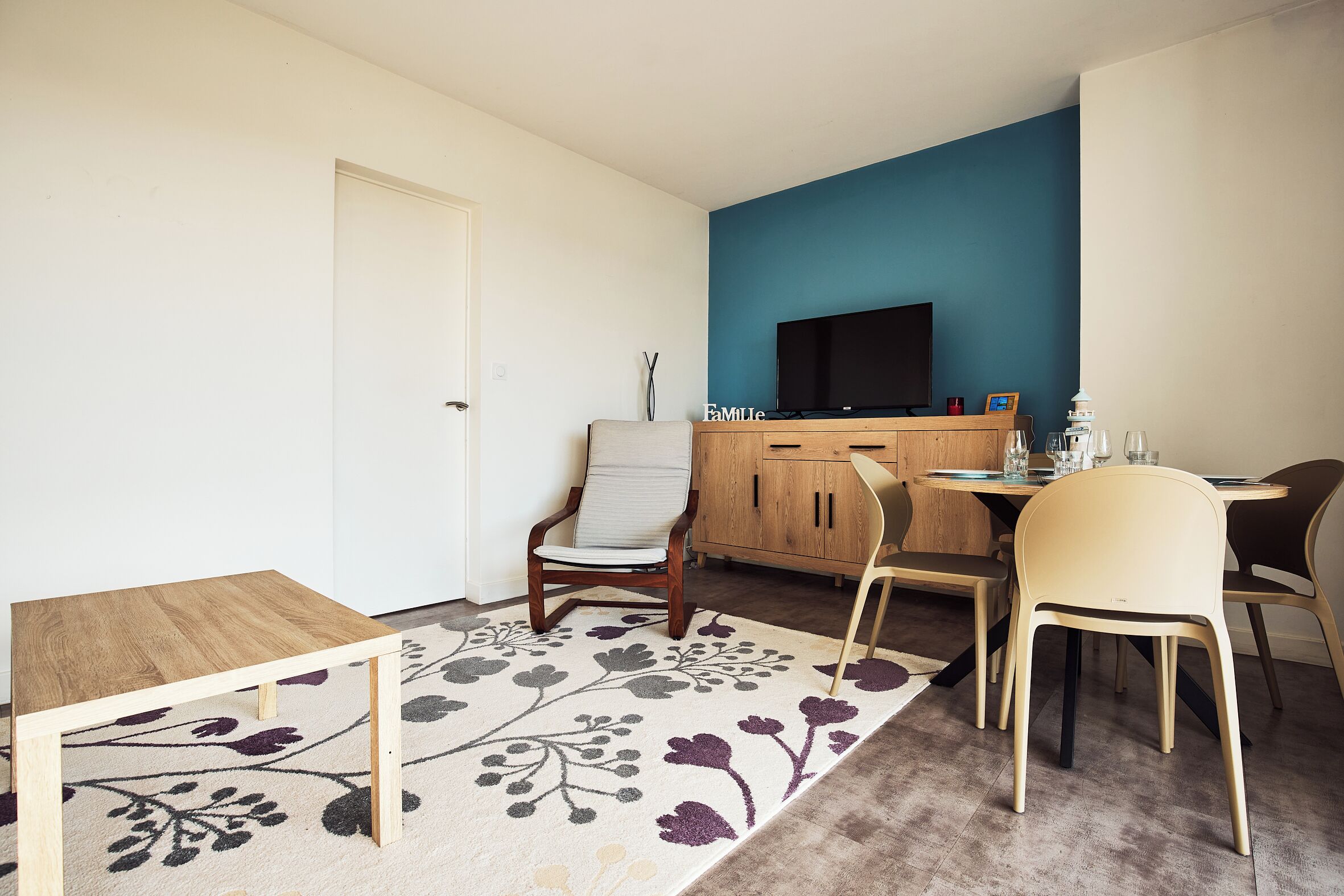 A small, well-lit room with a wooden table, a TV on a stand, and a dining area with chairs.