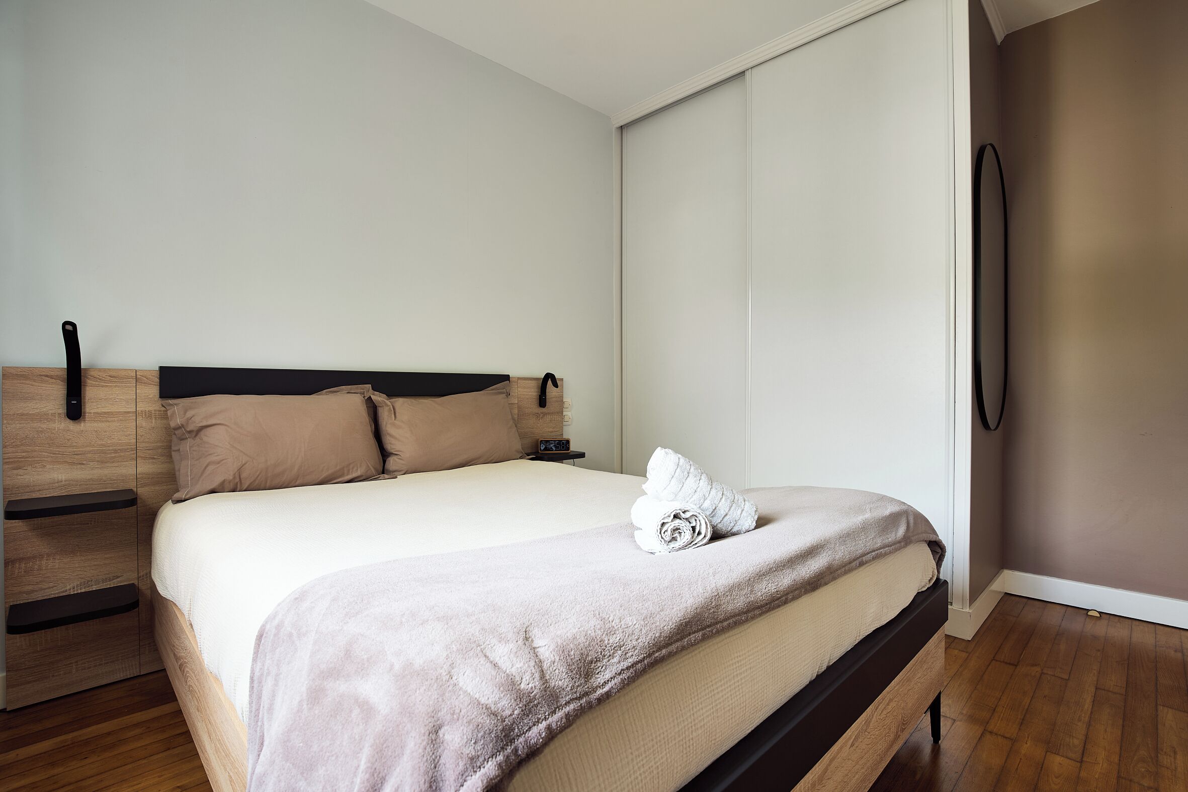 Una cama bien tendida con una manta color arena y una toalla enrollada, una mesita de noche de madera con una lámpara y un espejo circular fijado a la pared.