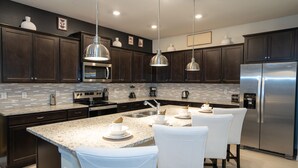 A modern kitchen with dark wood cabinets, stainless steel appliances, and a granite countertop. There are three white chairs around a small island. - Last Minute Disney! Private Pool, Hot Tub, Game Room & Themed Bedrooms. Book Now! #125797 (Orlando)