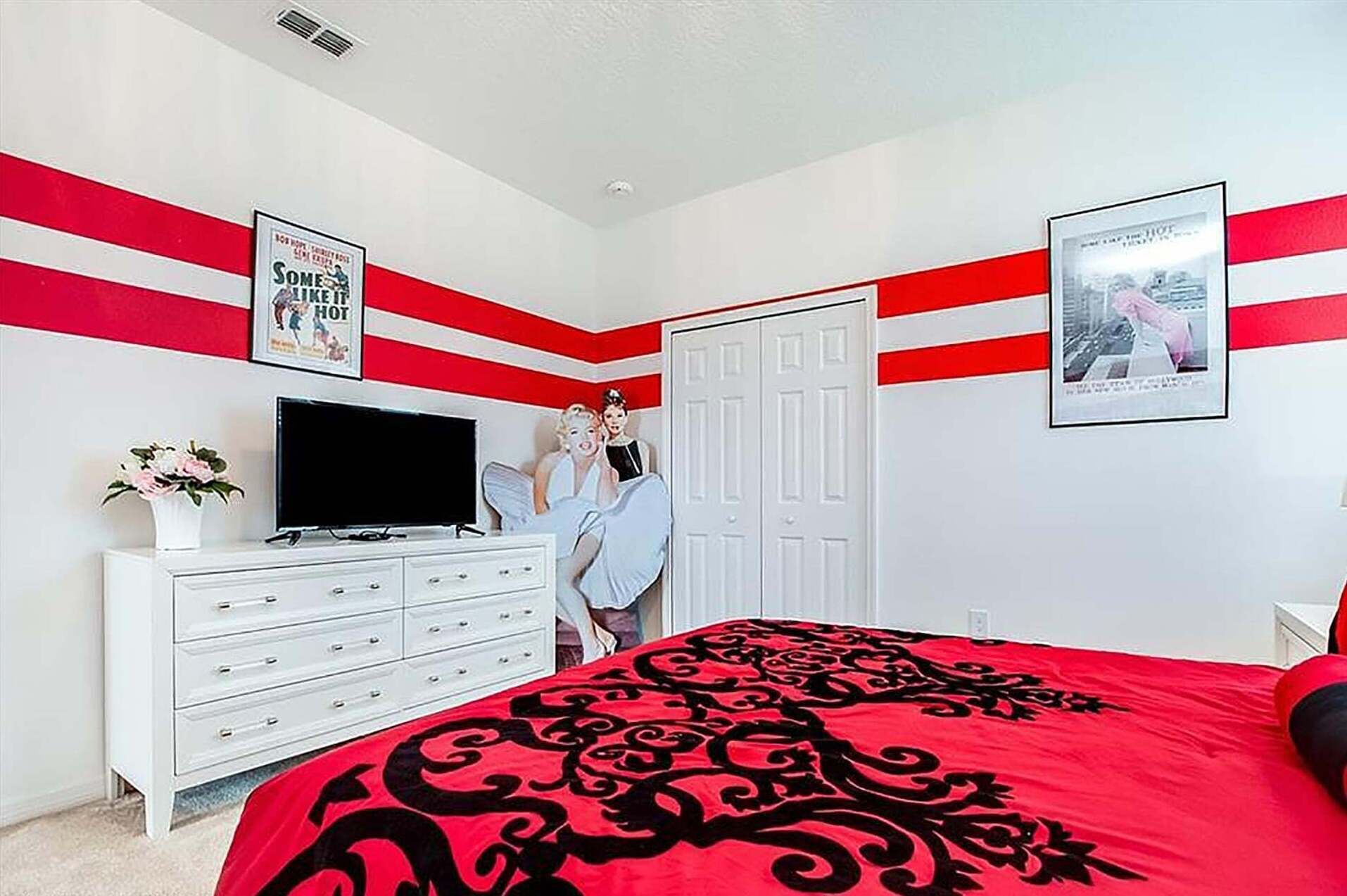 A bedroom with a red and white striped wall, a white dresser, a bed with a red and black patterned bedspread, and a television on the dresser.