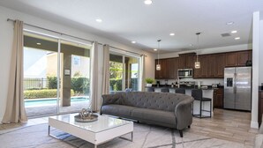 Una sala de estar moderna con sofá y mesa de centro, una cocina con electrodomésticos de acero inoxidable y un ventanal corredizo que da a una zona de piscina.