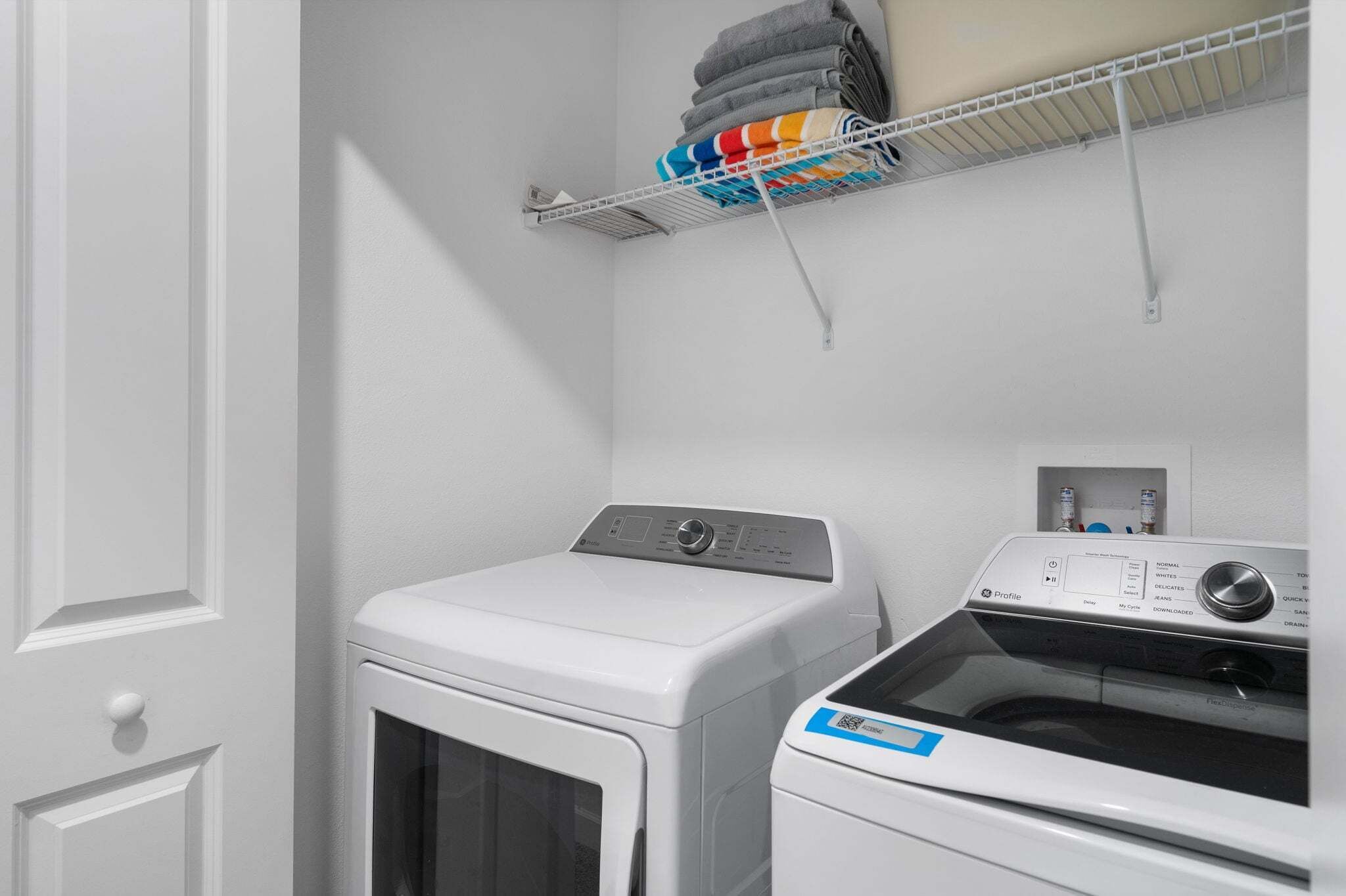 A laundry area with a front-loading washer and dryer, a metal shelf with folded towels, and a power outlet on the wall.