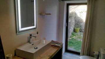 Een badkamer met een houten wastafel, een grote spiegel, een toilet en uitzicht op een stenen muur en groen buiten.