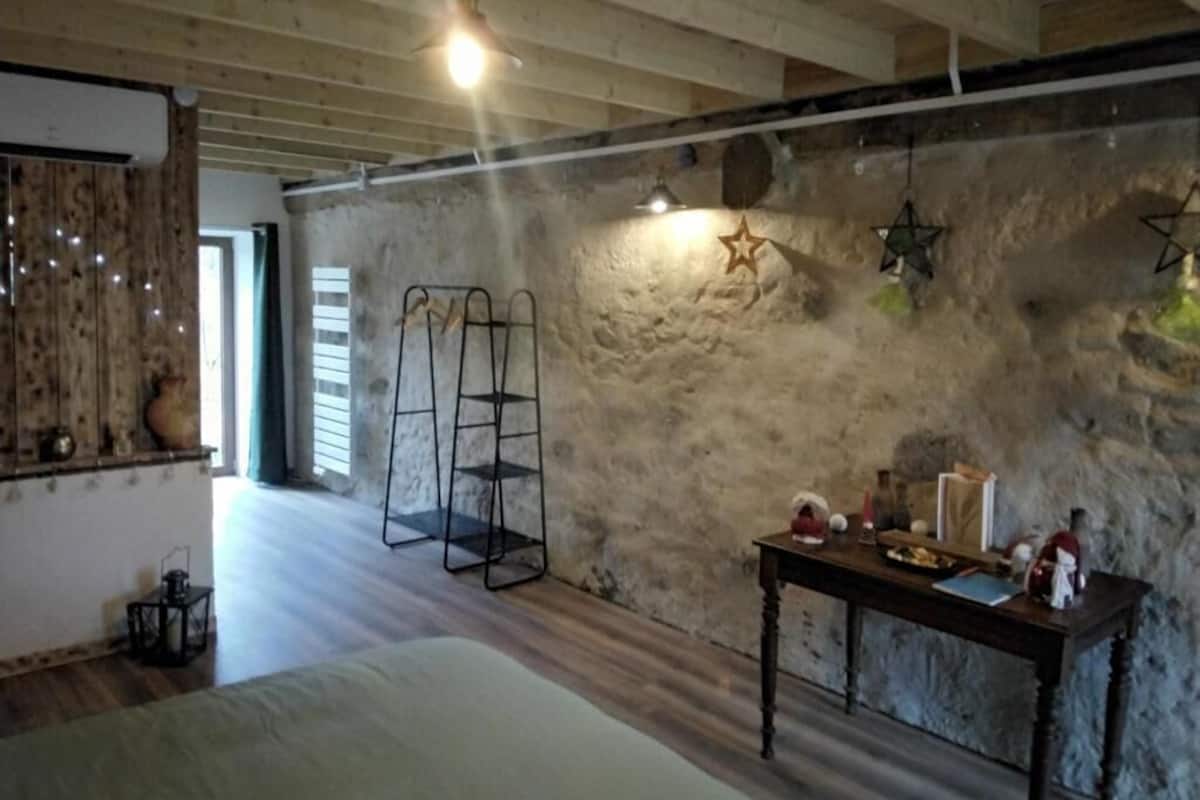 A rustic room with exposed wooden beams, a stone wall, a bed, a table with various items, and a ladder against the wall.