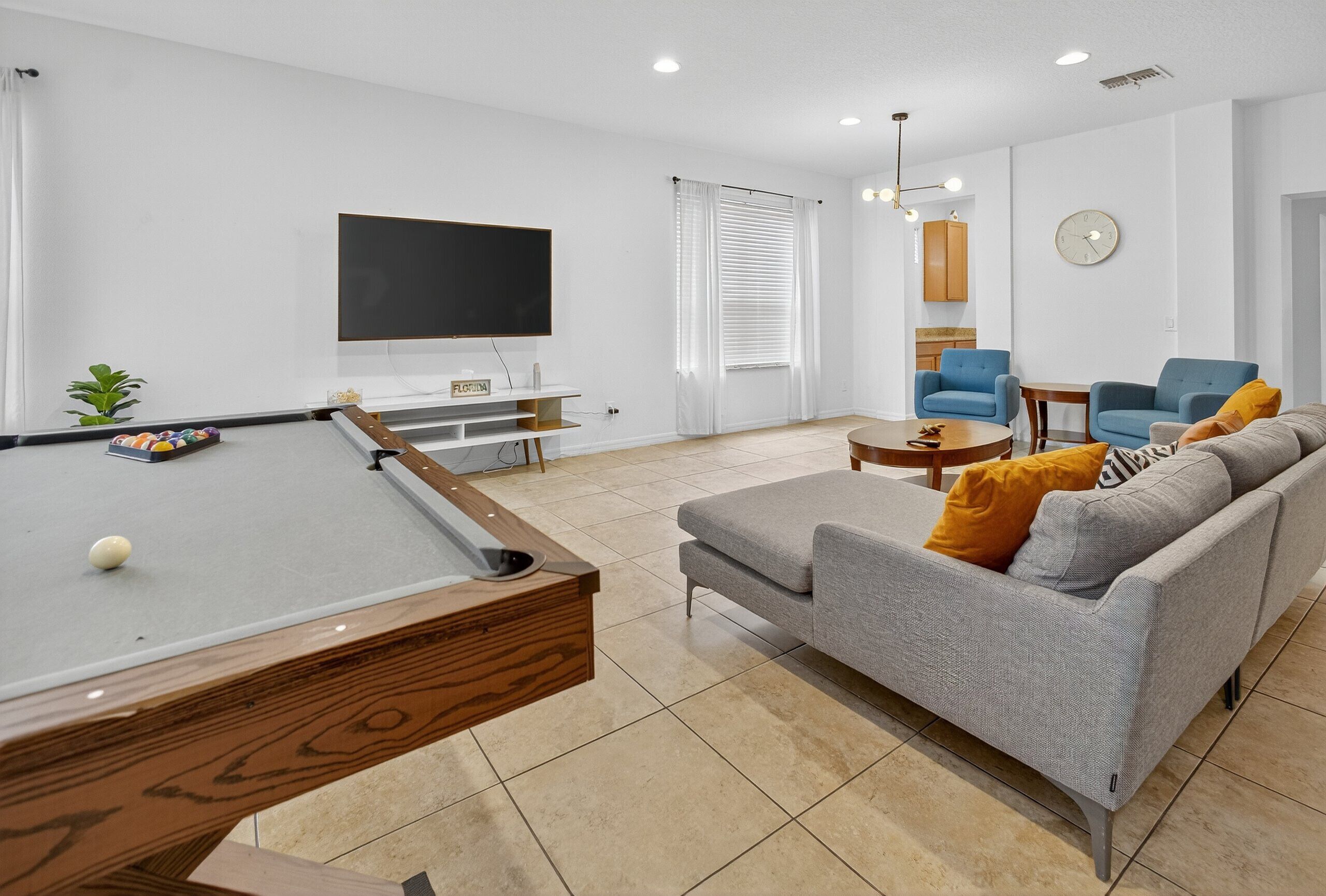 A modern living room with a pool table, a sofa with cushions, a television, and a clock on the wall.