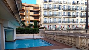 A swimming pool area with a tiled deck, a stone balustrade, and a view of multi-story buildings with balconies and windows. - Charming three-room beachfront apartment with stunning sea views in the heart of Menton (Menton)