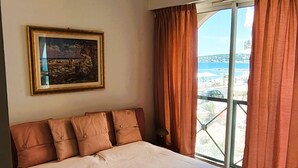 A bedroom with a large bed, a painting on the wall, and a view of the sea through the window. - Charming three-room beachfront apartment with stunning sea views in the heart of Menton (Menton)
