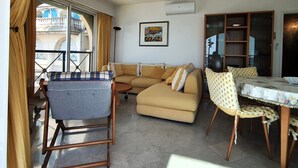 A living room with a sofa, a dining table with chairs, a standing chair, a window with curtains, and a wall-mounted air conditioner. - Charming three-room beachfront apartment with stunning sea views in the heart of Menton (Menton)