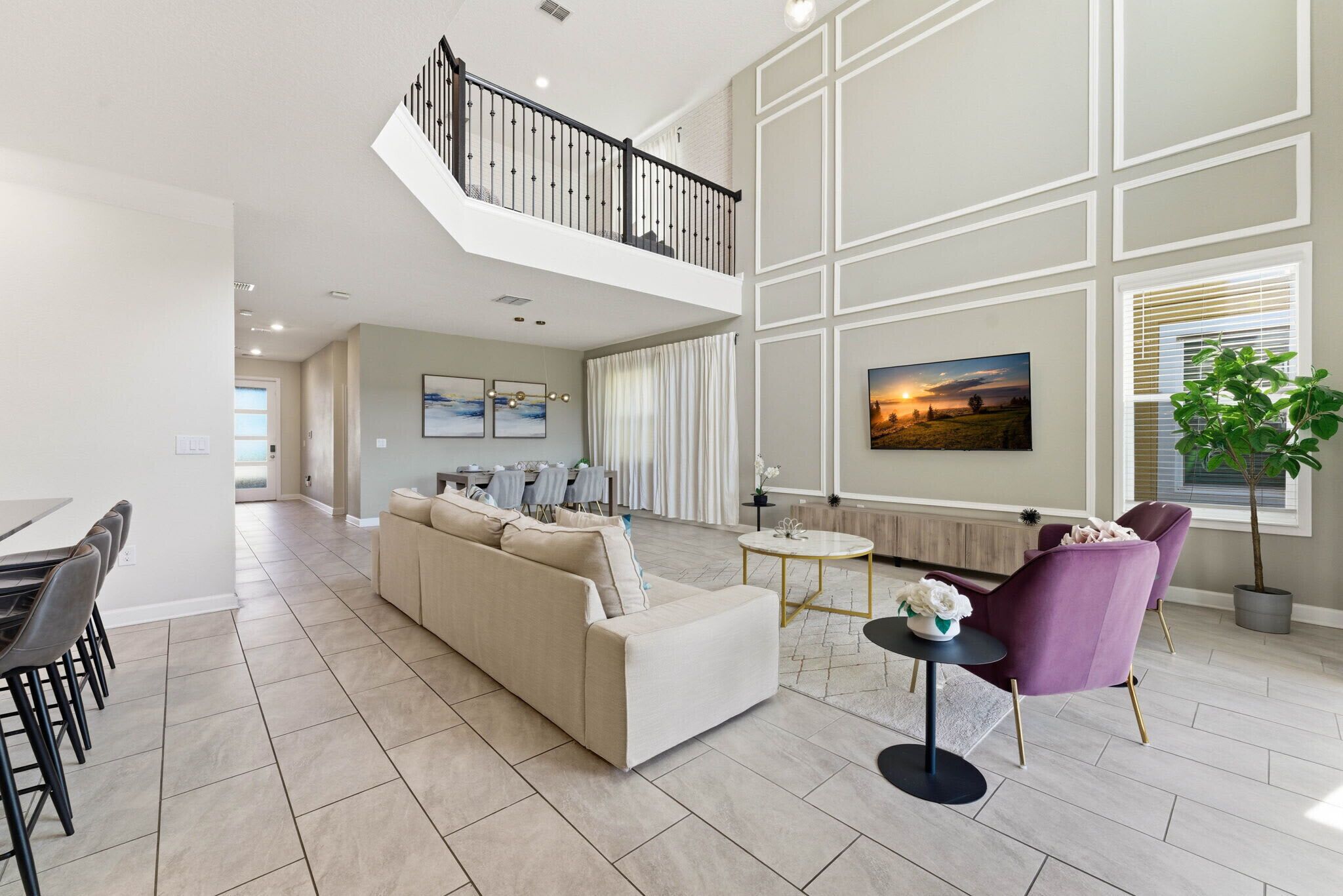 A spacious living area with a high ceiling, a balcony, a dining area, and a large window with blinds.