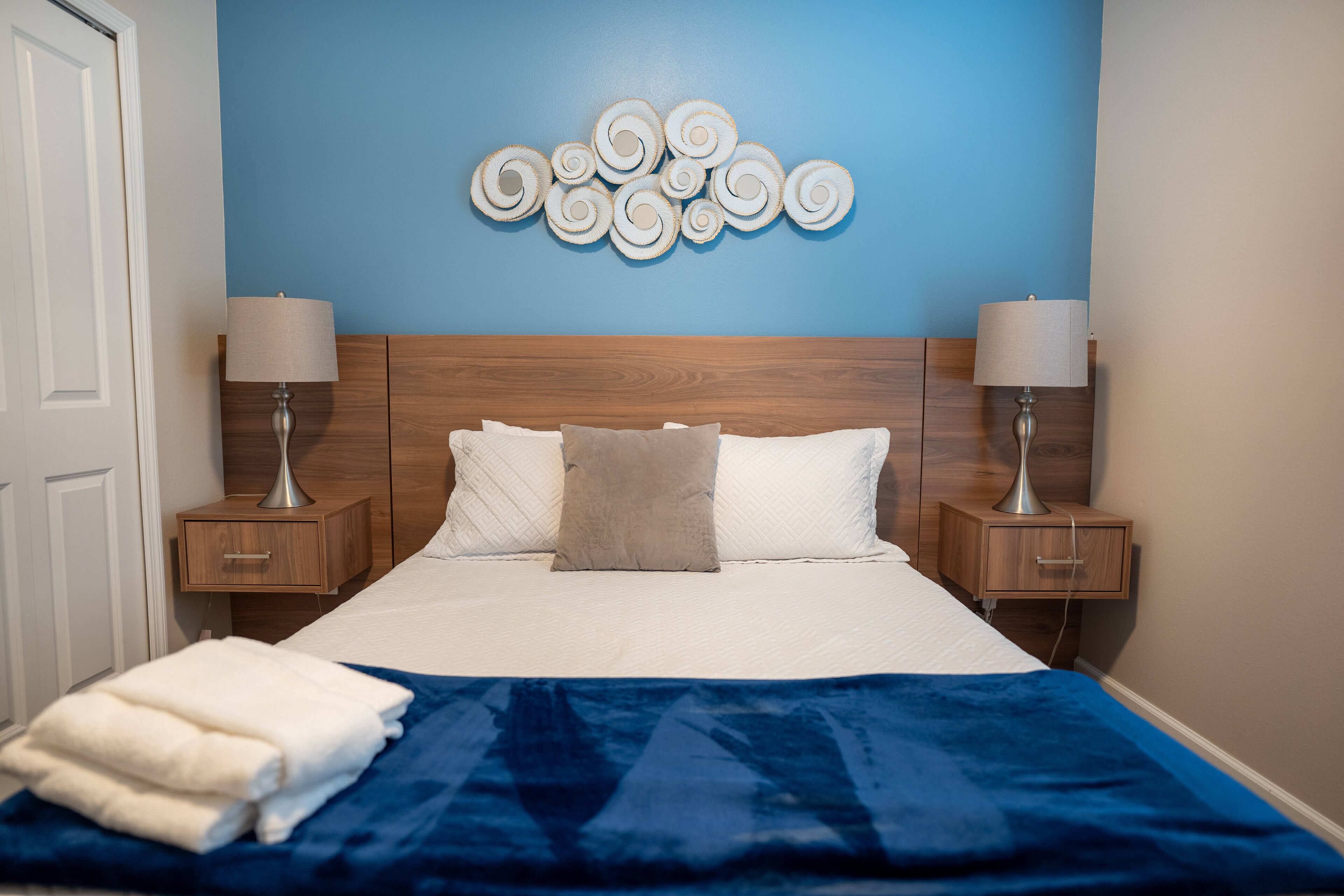Una cama bien hecha con un edredón azul, dos lámparas de noche y una obra de arte en la pared sobre la cama.