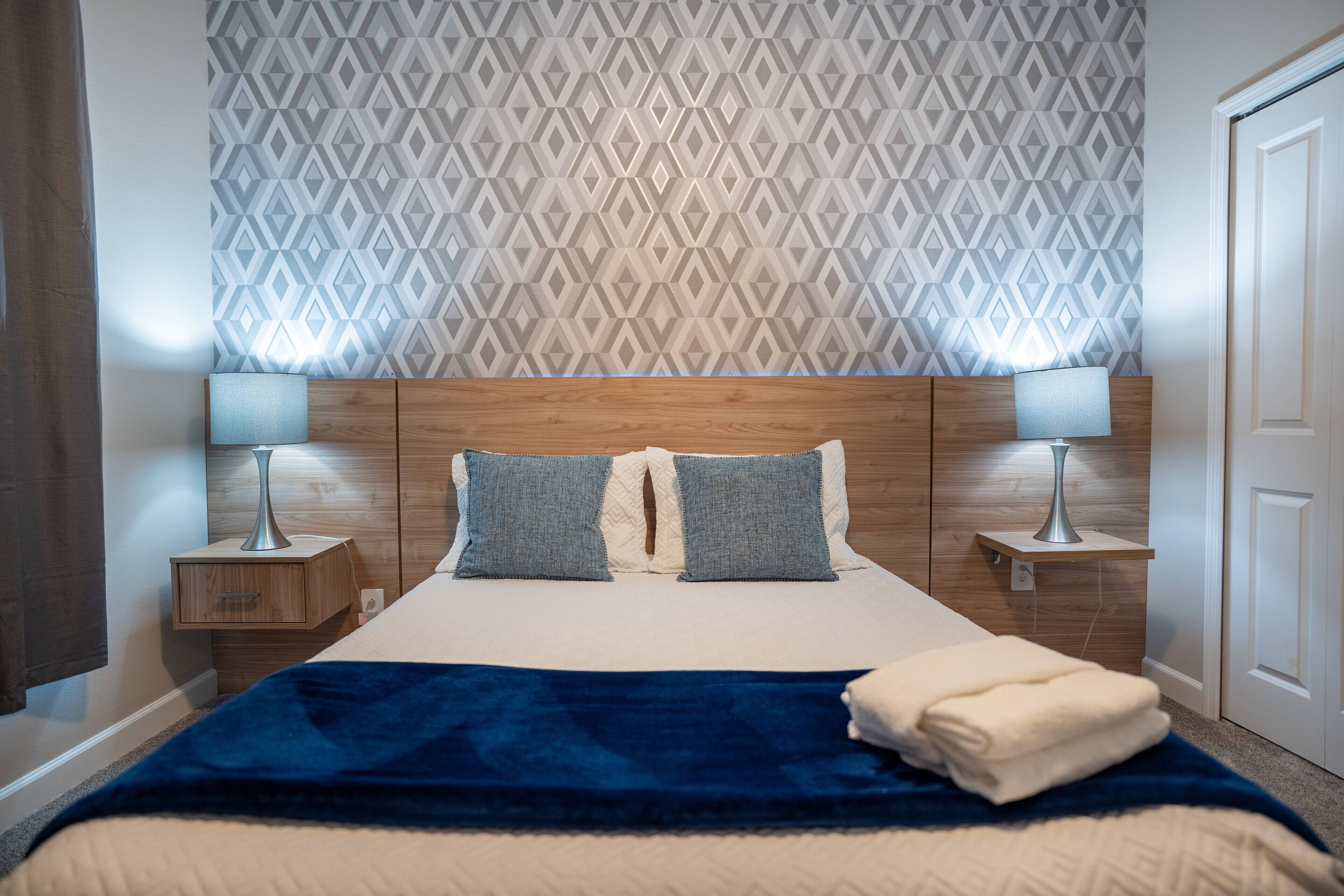 Ein ordentlich bezogenes Bett mit blauer Decke, zwei Kissen und einer Nachttischlampe auf jeder Seite.