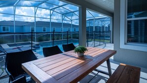 Un patio techado con una mesa y sillas de madera, una planta en maceta y vistas a una piscina y casas al fondo.