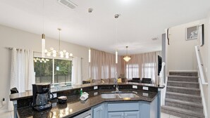 Uma cozinha moderna com bancada de granito, pia de aço inoxidável e lava-louças embutida. Há uma área de jantar com mesa e cadeiras, e uma escada que leva a outro nível.