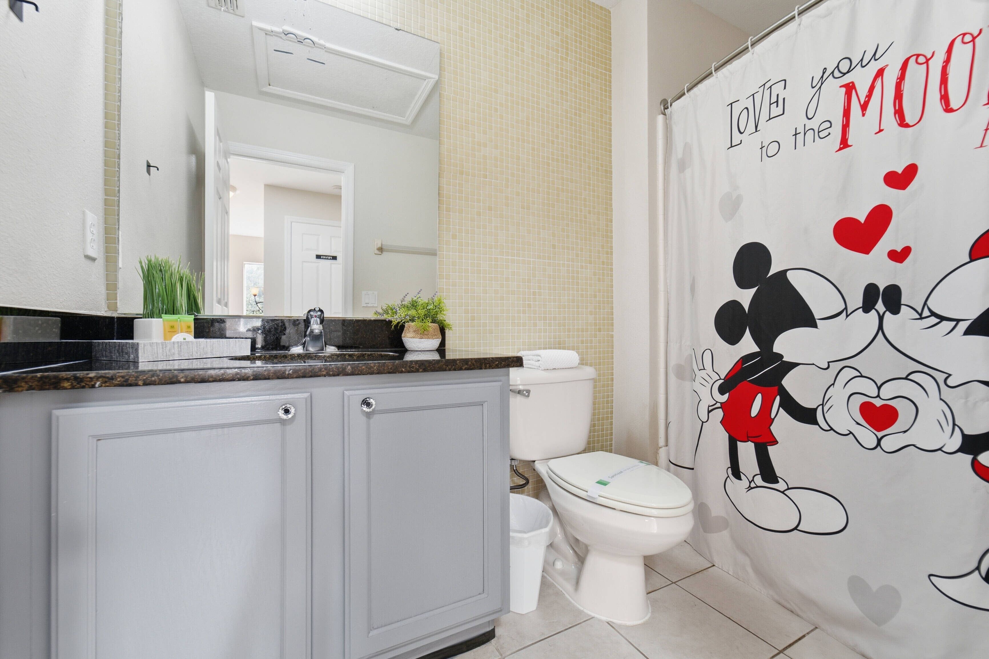 Une salle de bain avec un rideau de douche à l’effigie de Mickey Mouse, des toilettes blanches et un meuble gris doté d’un plan de travail.