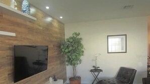 Ein modernes Wohnzimmer mit einem Flachbildfernseher an einer Holzwand, einer großen Topfpflanze, einem Ledersessel und einem kleinen Beistelltisch mit einer Vase.